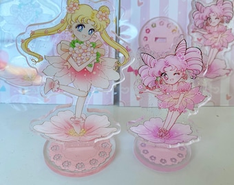 May include: Two pink and white acrylic character figurines of Sailor Moon characters. The figurines are standing on pink and white flower-shaped bases. The characters are holding flowers and have glitter accents.