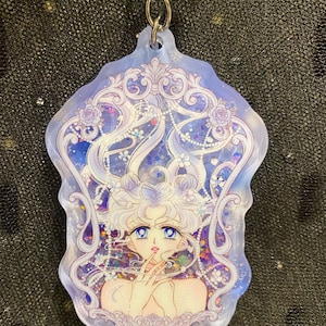 Op de afbeelding: Een transparante, iriserende sleutelhanger met een gedetailleerde illustratie van een jonge vrouw met lang, golvend haar, omringd door een decoratief frame met bloemmotieven. De sleutelhanger heeft een zilveren ketting.