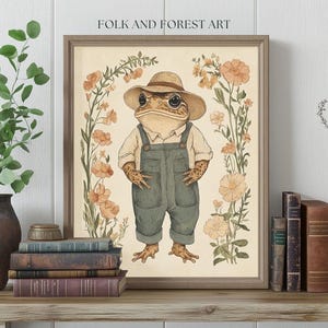 May include: A whimsical illustration of a frog wearing overalls and a straw hat, surrounded by delicate flowers in shades of pink and yellow. The artwork is framed in a light brown wood frame and features the text "Folk and Forest Art".