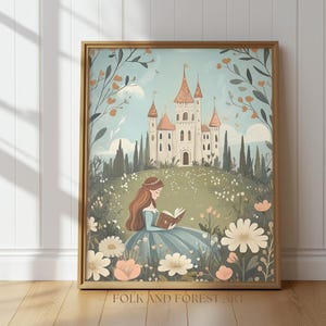 Puede incluir: Una impresión artística enmarcada representa una escena de cuento de hadas. Una mujer con un vestido azul lee un libro en un campo de flores, con un castillo al fondo. La impresión tiene una paleta de colores claros con el texto "FOLK AND FOREST ART" en la parte inferior.