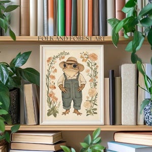 May include: A framed illustration of a frog wearing a hat and overalls, standing in a field of flowers. The illustration is in a watercolor style and has a vintage aesthetic. The text "FOLK AND FOREST ART" is at the top of the frame.