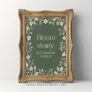 May include: A framed print with the text "Bloom slowly, but bloom surely" on a green background, surrounded by floral illustrations. The frame is ornate with a gold finish. The words "FOLK AND FOREST ART" are at the bottom.