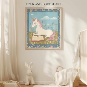 May include: A framed art print featuring a white unicorn with a pink mane and tail, reading a book in a field of flowers. The artwork has a blue border and is labeled "FOLK AND FOREST ART" at the top.