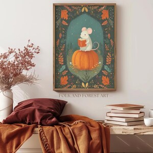 Op de afbeelding: Een ingelijste kunstprint toont een witte muis die een boek leest op een oranje pompoen, tegen een achtergrond van herfstbladeren. Het kunstwerk bevat de tekst "FOLK AND FOREST ART" en is ingelijst in een houten lijst.