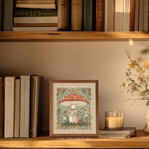 Puede incluir: Una impresi&oacute;n art&iacute;stica enmarcada que presenta un rat&oacute;n bajo una seta roja y blanca, rodeado de flores y vegetaci&oacute;n. La impresi&oacute;n se exhibe en una estanter&iacute;a de madera con libros y una vela encendida. El texto "FOLK AND FOREST ART" est&aacute; abajo.