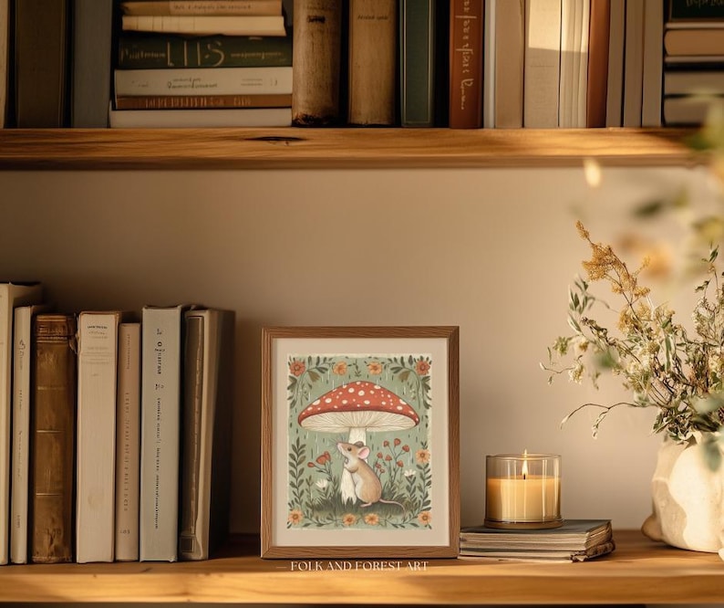 Puede incluir: Una impresi&oacute;n art&iacute;stica enmarcada que presenta un rat&oacute;n bajo una seta roja y blanca, rodeado de flores y vegetaci&oacute;n. La impresi&oacute;n se exhibe en una estanter&iacute;a de madera con libros y una vela encendida. El texto "FOLK AND FOREST ART" est&aacute; abajo.