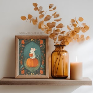 Op de afbeelding: Een ingelijste kunstafdruk met een illustratie van een muis die een boek leest op een pompoen. De print heeft een teal achtergrond met bloemmotieven. Een kaars en een vaas met gedroogde takken staan ook op de plank.