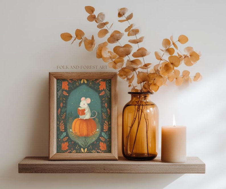 Op de afbeelding: Een ingelijste kunstafdruk met een illustratie van een muis die een boek leest op een pompoen. De print heeft een teal achtergrond met bloemmotieven. Een kaars en een vaas met gedroogde takken staan ook op de plank.