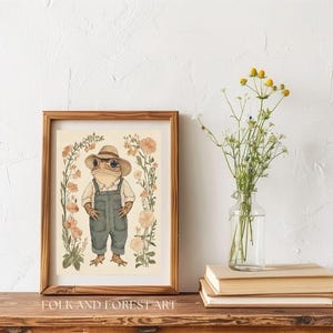 May include: A watercolor illustration of a frog wearing a straw hat and overalls standing in a field of flowers. The illustration is framed in a brown wood frame and the text "FOLK AND FOREST ART" is printed below the frame.