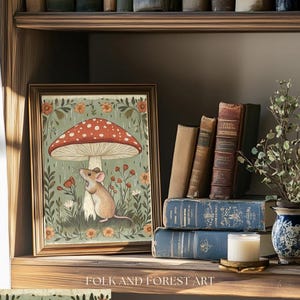 Puede incluir: Una impresi&oacute;n art&iacute;stica enmarcada que presenta un rat&oacute;n bajo una seta roja y blanca, rodeado de flores. La obra de arte se exhibe en una estanter&iacute;a de madera con libros antiguos, una vela y un jarr&oacute;n decorativo. La impresi&oacute;n est&aacute; etiquetada como "Folk and Forest Art".