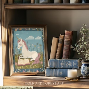 May include: A framed print of a unicorn reading a book, set on a wooden shelf with antique books and a small vase of greenery. The unicorn has a white body, pink mane and tail, and a golden horn. The print is surrounded by a gold frame.