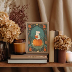 Op de afbeelding: Een ingelijste kunstdruk met een muis die een boek leest op een pompoen. De print staat op een houten plank met boeken, een kaars en droogbloemen. De tekst "FOLK AND FOREST ART" is zichtbaar. Herfsttinten.