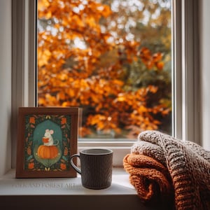 Op de afbeelding: Een ingelijste prent van een muis die op een pompoen leest, een bruine mok en een opgevouwen deken liggen op een vensterbank. De prent heeft een turquoise achtergrond met oranje en groene accenten. De mok is donkerbruin. De deken is oranje en beige.