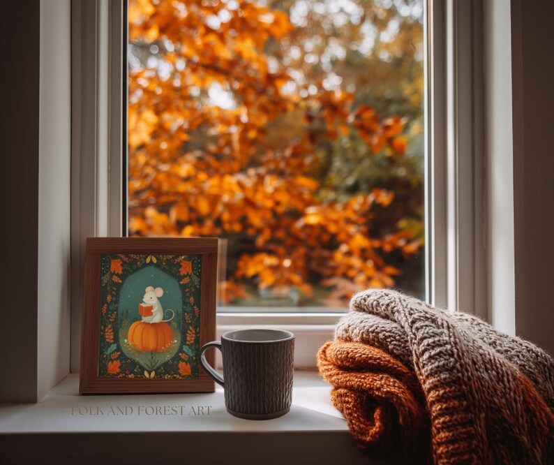 Op de afbeelding: Een ingelijste prent van een muis die op een pompoen leest, een bruine mok en een opgevouwen deken liggen op een vensterbank. De prent heeft een turquoise achtergrond met oranje en groene accenten. De mok is donkerbruin. De deken is oranje en beige.