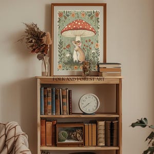 Puede incluir: Una impresi&oacute;n art&iacute;stica enmarcada que presenta una ilustraci&oacute;n de un rat&oacute;n bajo una seta roja y blanca. La obra de arte se encuentra sobre un fondo verde y floral. La impresi&oacute;n se exhibe en una estanter&iacute;a de madera con libros, un reloj y objetos decorativos.