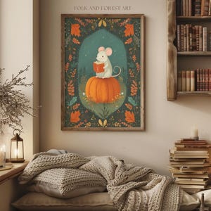 Op de afbeelding: Een ingelijste kunstprint met een grillige illustratie. Een witte muis leest een boek terwijl hij op een grote oranje pompoen zit. Het kunstwerk is geplaatst tegen een teal achtergrond met herfstbladeren en de tekst "FOLK AND FOREST ART" bovenaan.