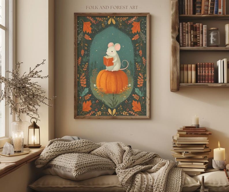 Op de afbeelding: Een ingelijste kunstprint met een grillige illustratie. Een witte muis leest een boek terwijl hij op een grote oranje pompoen zit. Het kunstwerk is geplaatst tegen een teal achtergrond met herfstbladeren en de tekst "FOLK AND FOREST ART" bovenaan.