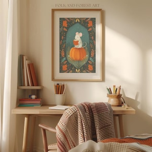 Op de afbeelding: Ingelijste kunstdruk met een witte muis die een boek leest op een oranje pompoen. De achtergrond is donkerblauwgroen met bloemen en bladeren. De lijst is lichtbruin. Een charmant kunstwerk.