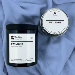 May include: A black candle jar with a black lid and a white label that reads "Two Palms Candle Co. Twilight Amber, Berries, Woods 15.0 oz 425 g 100% Natural Soy Wax Hand-Poured in San Diego, CA Small Batch 70 Hr Burn Time". The candle jar is sitting on a blue fabric surface.