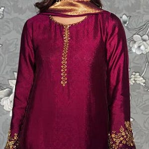 May include: A long-sleeved, burgundy-colored dress with gold embroidery on the cuffs and bodice. The dress features gold accents on the hem and is paired with matching pants. The outfit is suitable for special occasions.