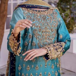 May include: A teal-colored, long-sleeved garment with intricate gold embroidery. The embroidery covers the bodice, sleeves, and cuffs. The cuffs and neckline are trimmed with gold and navy blue accents. The garment has a flowing, elegant design.