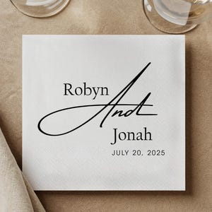 以下が含まれることがあります： 黒い文字が入った白い正方形のナプキン。「Robyn」と「Jonah」という名前が、中央に様式化された「And」とともに表示されています。日付「JULY 20, 2025」が下に印刷されています。ワイングラスが2つ見えます。