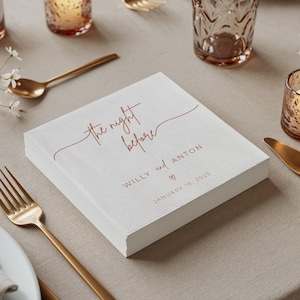 May include: A stack of white paper napkins with the text "the night before" in copper-colored script. Below, the names "Willy and Anton" are printed, along with the date "January 16, 2025". The napkins are on a beige tablecloth, with gold cutlery and candle holders.