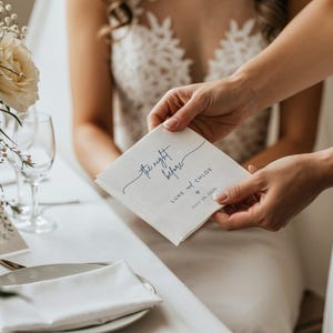 May include: A square, off-white wedding napkin with the text "the night before" in a flowing, dark blue script. Below, it reads "Luke and Chloe" and the wedding date, July 28, 2026. The napkin is held by two hands.