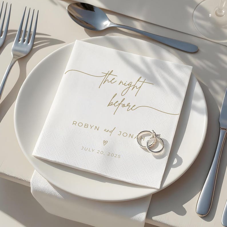 May include: A white napkin with gold script reading "the night before" and names, a heart, and a date. Two silver rings rest on the napkin, which is on a white plate with silverware. A spoon and fork are visible.