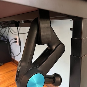 May include: A black and blue massage gun hanging from a desk with a black 3D printed holder. The gun has a blue circular button and a black handle.