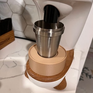 May include: A white and beige electric ice cream maker with a stainless steel cup attached to the bottom. The ice cream maker has a wooden base.