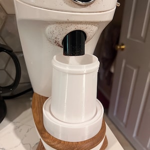 May include: A white coffee grinder with a wooden base. The grinder has a black chute and a white portafilter holder. The grinder is labeled "NICHE" in black letters on a white circle.