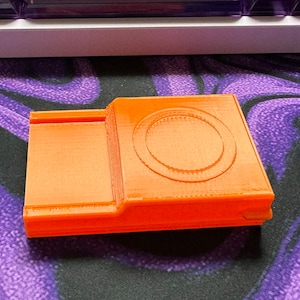 Pode incluir: Um objeto laranja impresso em 3D com uma indentação circular no topo. O objeto tem uma superfície lisa e texturizada.