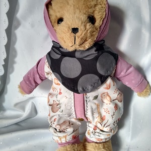 以下が含まれることがあります： ピンクのフード付きロンパースと黒と白のドット柄のスタイをつけた茶色のテディベア。ロンパースは白い背景に動物と植物の模様が描かれています。