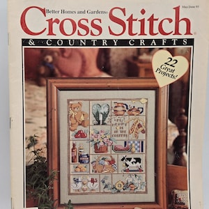 May include: The cover of a "Better Homes and Gardens Cross Stitch & Country Crafts" magazine from May/June 1993. The cover features a framed cross-stitch sampler with various country-themed motifs, including a teddy bear, flowers, and a cow. A heart-shaped sticker reads "22 Great Projects!"