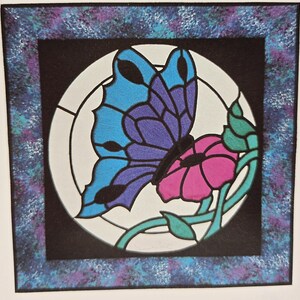 May include: A vibrant art print featuring a butterfly and a flower. The butterfly's wings are blue and purple, and the flower is pink. The artwork is set against a black background within a white circle, framed by a speckled blue and purple border.
