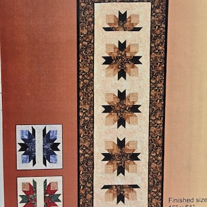 May include: A quilted table runner with a cream-coloured central panel featuring repeating starburst designs in shades of brown and black. The border is a dark floral pattern. Finished sizes are 40.6cm x 129.5cm and 50.8cm x 142.2cm.