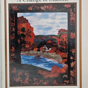 May include: A pattern booklet titled Joan's Own Creations A Change of Seasons Applique Series 120. The cover features a landscape illustration of a white house by a blue river surrounded by autumn trees with red and orange foliage under a blue sky.