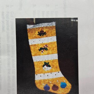 May include: A Christmas pocket stocking with the text CHRISTMAS POCKET STOCKING at the top. The stocking features alternating horizontal yellow and white stripes, decorated with small embroidered holiday icons including a candle, candy cane, and snowman.
