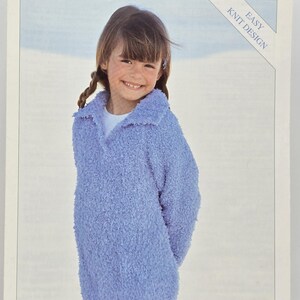 Pode incluir: Uma criança vestindo um suéter de malha grossa azul claro com gola. O suéter faz parte de um padrão "Easy Knit Design" da Sirdar, rotulado como "Snowflake". A imagem apresenta uma criança sorrindo e o texto "4170".