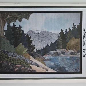 Op de afbeelding: Een rechthoekig patroonboekje getiteld Joan's Own Creations Mountain Vista Appliqué Series 139. De omslag bevat een landschapsillustratie van een besneeuwde berg, donkergroene dennenbomen, een rotsachtige kustlijn en een blauw meer.