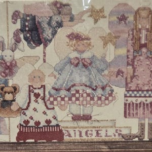 May include: Cross-stitch artwork depicting angels with wings, dressed in pastel colours. The angels are surrounded by stars and toys, with the word "ANGELS" at the bottom. The artwork is framed in a wooden frame.
