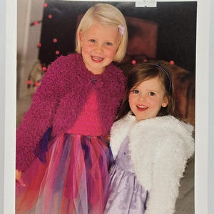 May include: Two young girls smiling, wearing matching outfits. One wears a pink dress and a magenta fuzzy jacket, the other a lavender dress and a white fuzzy shawl. The image has text that reads "SNOWFLAKE GLACIER" and "SIRDAR".