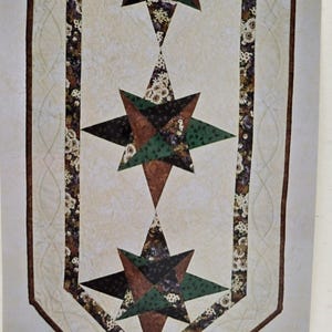 May include: A quilted table runner featuring three starburst designs in shades of brown, green, and black. The stars are set against a cream-colored background with floral patterned borders and a brown border.