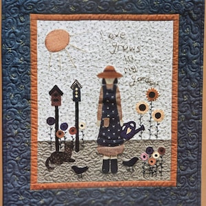 May include: A quilted wall hanging with a garden theme. The artwork features a figure in a hat and apron watering sunflowers, with the text "Love grows in my garden." A cat, birds, and birdhouses are also depicted.
