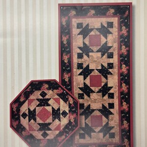 May include: A rectangular table runner and an octagonal mat featuring a patchwork design with red, black, and tan fabric. The patterns include floral motifs and geometric shapes. The text Perkins Dry Goods is printed at the top of the image.
