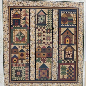 Op de afbeelding: Een vierkante quilt met een patchworkontwerp van diverse vogelhuisjes, geometrische sterpatronen en boommotieven in marineblauw, bordeauxrood, mosterdgeel en bosgroen, omlijst door een bloemenrand met blauwe en bruine ranken.