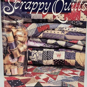 May include: The cover of a book titled "Simply Scrappy Quilts" showing a stack of patchwork quilts in various colours and a jar filled with thread spools. The cover has a red, white, and blue patchwork design, with the author's name, Nancy J. Martin.