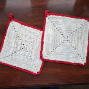 May include: Two square, cream-coloured crochet potholders with a red border and a loop for hanging. Each potholder features a decorative stitched design in the centre. The potholders are displayed on a dark brown wooden surface.