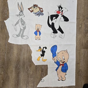 Puede incluir: Tela blanca con personajes de dibujos animados Bugs Bunny, Taz, Silvestre, Porky Pig y Pato Lucas. Los personajes están en sus poses y atuendos clásicos. La tela está cortada en formas irregulares.